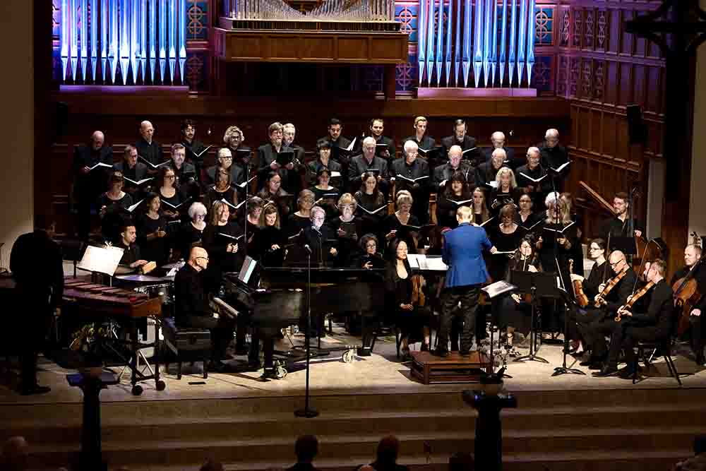 Large choir sings on risers behind a conductor leading an orchestra on stage, with organ pipes and wood-paneled walls in the background.
