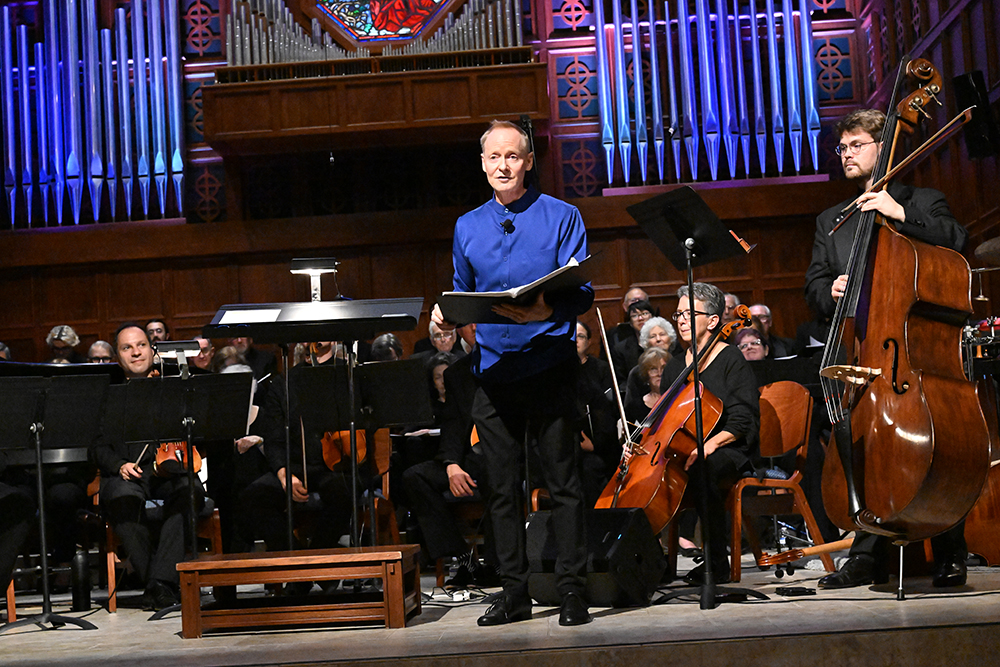 Man in a blue shirt stands at a music stand speaking or singing during a live performance, with an orchestra seated behind him and a double bass player to the right on a concert hall stage