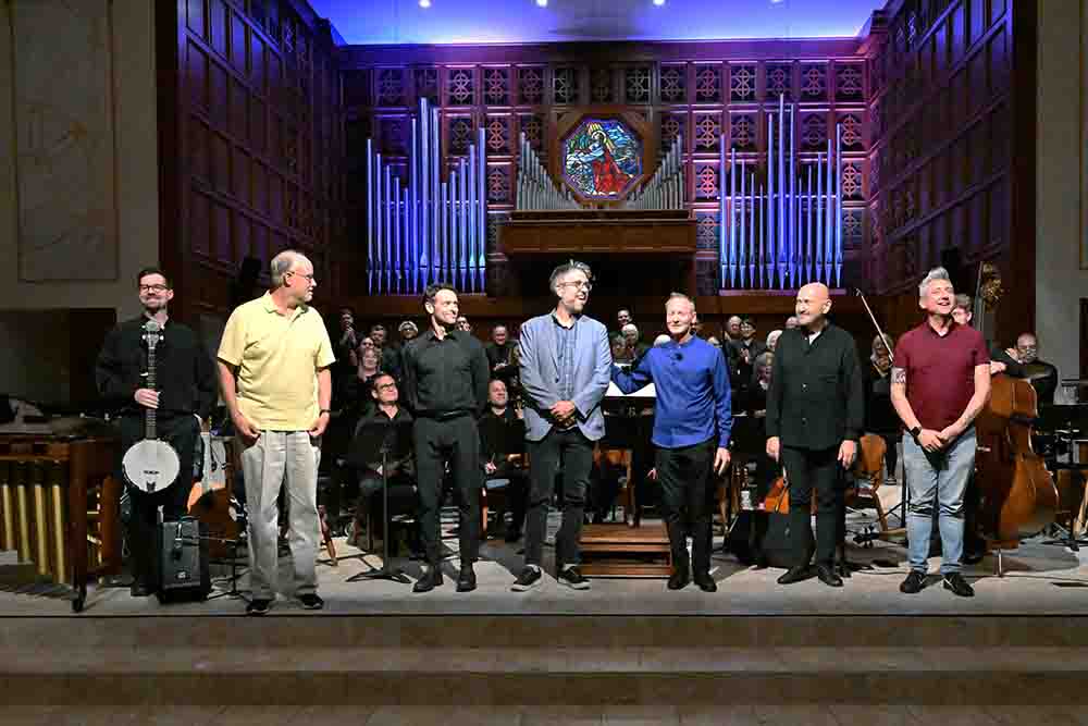 Seven performers stand across a stage in front of an orchestra and choir, facing the audience beneath large organ pipes in a concert hall.