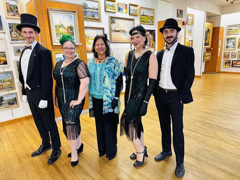 Five adults dressed in 1920s-inspired formal attire, including flapper dresses, gloves, and top hats, stand together smiling inside an art gallery with framed paintings on the walls.