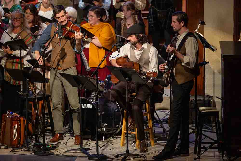 Small group of musicians plays violin, guitar, and banjo on stage, with additional performers and instruments visible behind them.