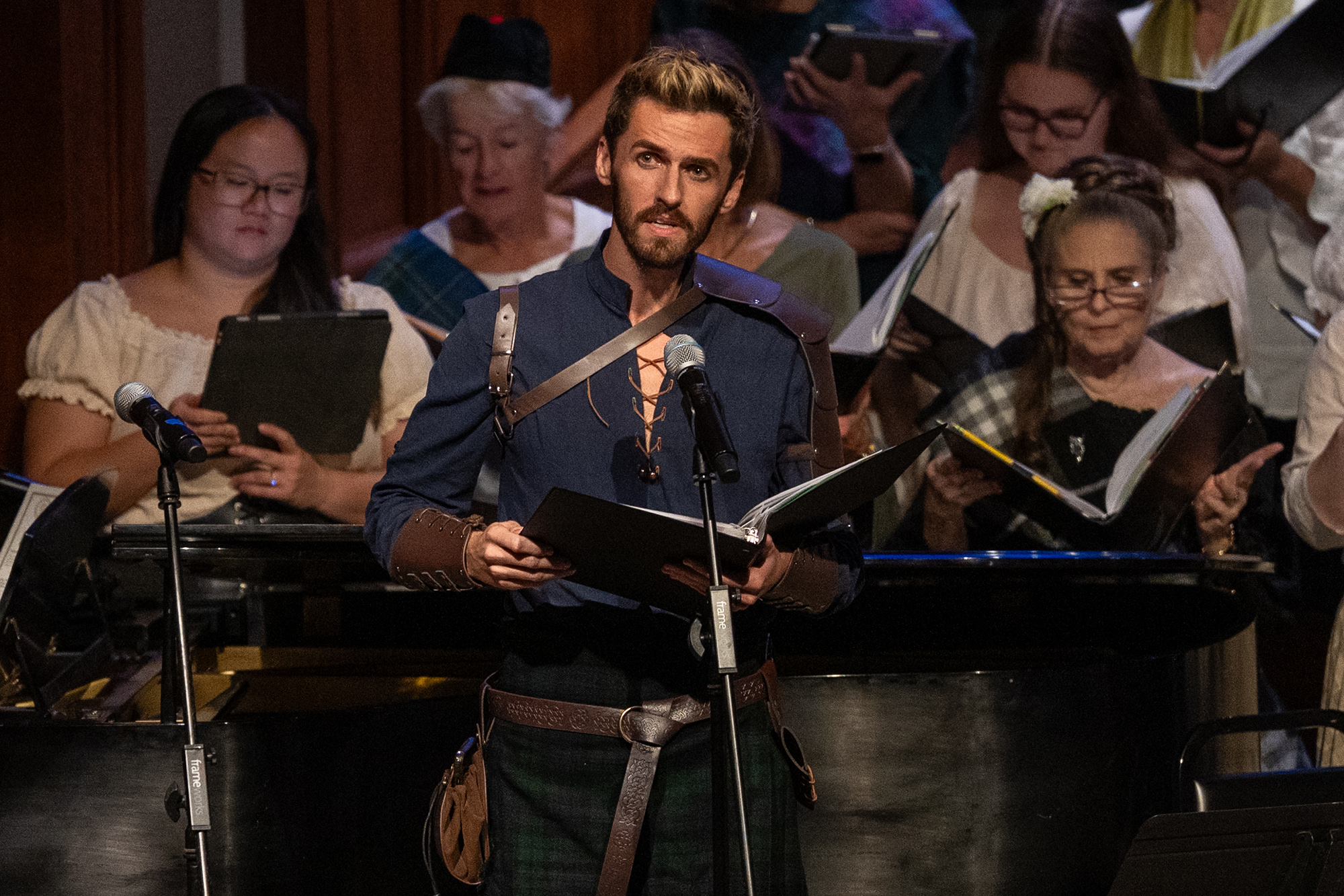 Male soloist in a medieval-style costume sings into a microphone while holding a music binder, with a choir standing behind him.